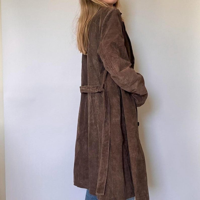May include: A long, brown suede coat with a belt. The coat has a classic design with a notched collar and a button closure. The coat is knee-length and has a relaxed fit.
