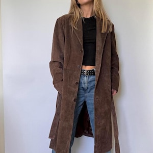 May include: A long, brown suede coat with a button-up front and a matching belt. The coat is knee-length and has a classic, tailored design. The coat is worn over a black top and blue jeans.