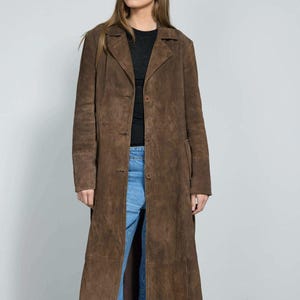 May include: A long, brown suede coat with a button-up front and notched lapels. The coat is knee-length and has a classic, tailored design. The model is wearing blue jeans and a black top.