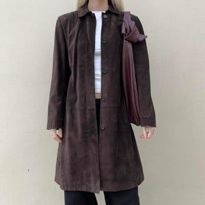 May include: A long, dark brown suede coat with a button-up front and a collared neckline. The coat is worn over a white top and black pants. A dark purple tote bag is carried over the shoulder.