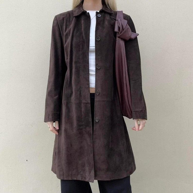 May include: A long, dark brown suede coat with a button-up front and a collared neckline. The coat is worn over a white top and black pants. A dark purple tote bag is carried over the shoulder.