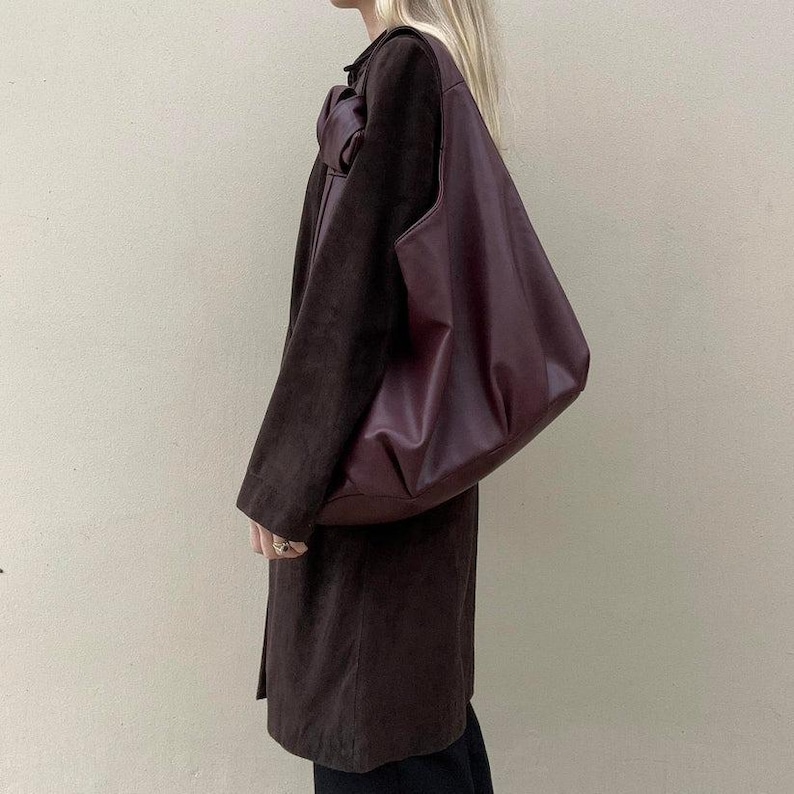 May include: A dark burgundy leather tote bag with a single shoulder strap. The bag is held against a person wearing a long, dark brown suede coat. The coat has a large bow detail at the shoulder.