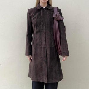 May include: A brown suede coat with a collared neckline and long sleeves. A dark purple tote bag is draped over the shoulder. The coat has a button closure and a tailored fit, suitable for fall or winter wear.