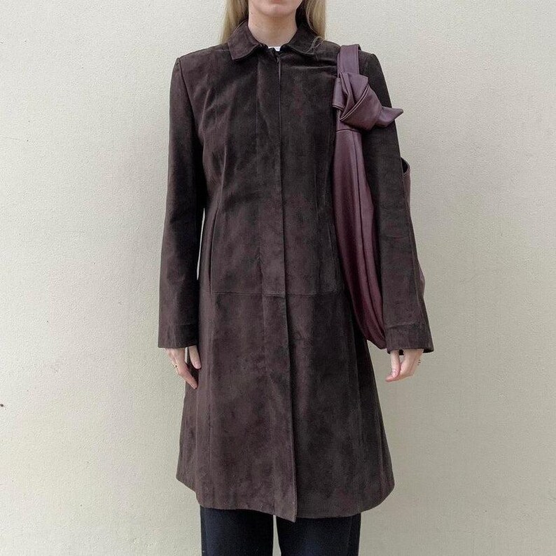May include: A brown suede coat with a collared neckline and long sleeves. A dark purple tote bag is draped over the shoulder. The coat has a button closure and a tailored fit, suitable for fall or winter wear.