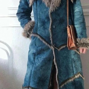 May include: A long, teal suede coat with faux fur trim on the collar, cuffs, and edges. The coat features a button-down front and a small brown leather purse with a long strap. The coat is a vintage style.