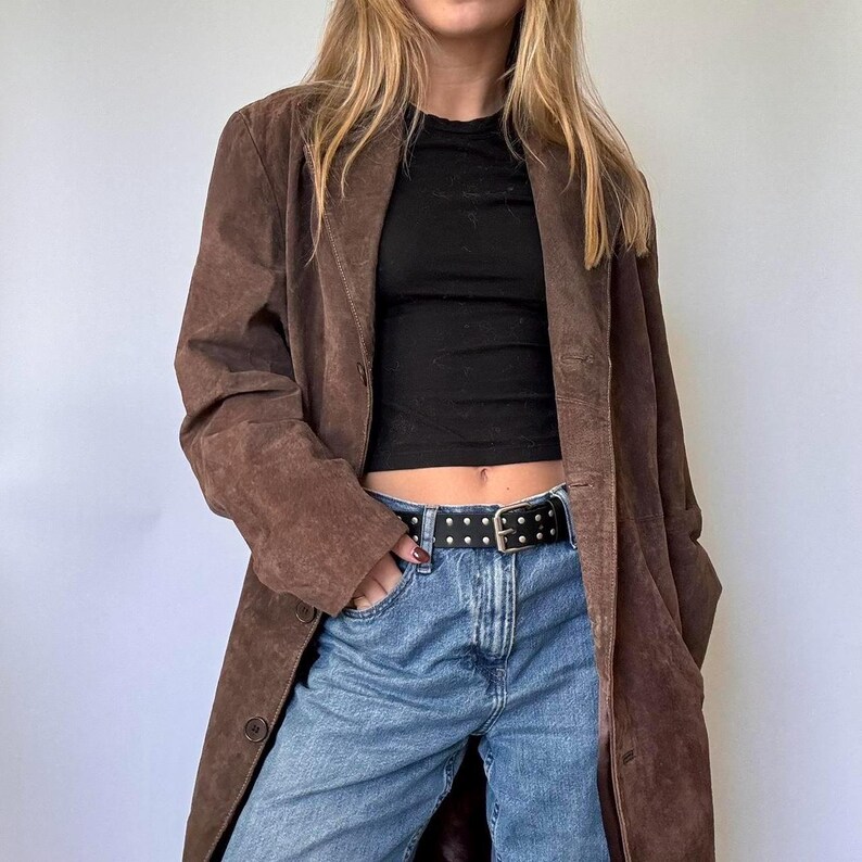 May include: A long, brown suede coat with a button closure. The coat is worn over a black top and blue jeans. A black belt with silver studs is worn around the waist.
