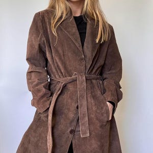 May include: A brown suede trench coat with a belted waist and button fastening. The coat has a classic lapel collar and long sleeves. The coat is knee-length and has pockets.