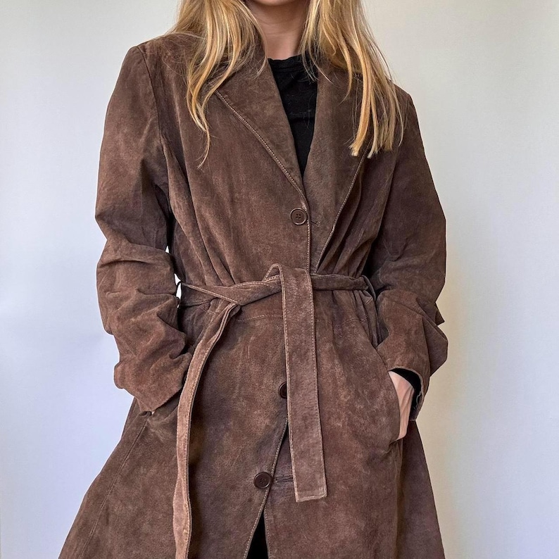 May include: A brown suede trench coat with a belted waist and button fastening. The coat has a classic lapel collar and long sleeves. The coat is knee-length and has pockets.