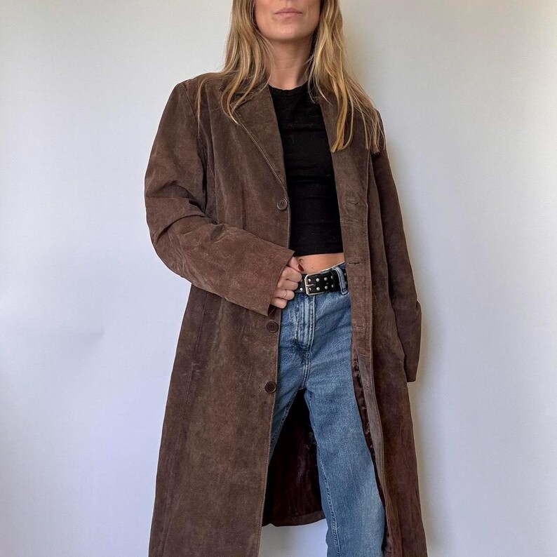 May include: A long, brown suede coat with a button closure. The coat has a classic lapel collar and long sleeves. The coat is worn over a black top and blue jeans.