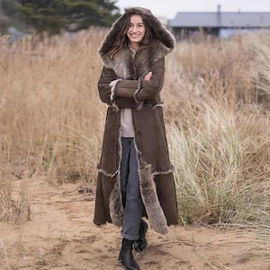 May include: A long, brown coat with a fur-lined hood and trim. The coat is buttoned up and appears to be made of suede or a similar material. The person is wearing gray pants and black boots. The coat is a winter outerwear garment.