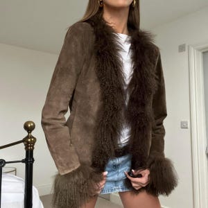 Puede incluir: Chaqueta de ante marr&oacute;n con ribete de piel sint&eacute;tica marr&oacute;n oscuro alrededor del cuello y los pu&ntilde;os. La chaqueta tiene un corte cl&aacute;sico y se usa sobre una camiseta blanca y una falda vaquera. Estilo vintage.