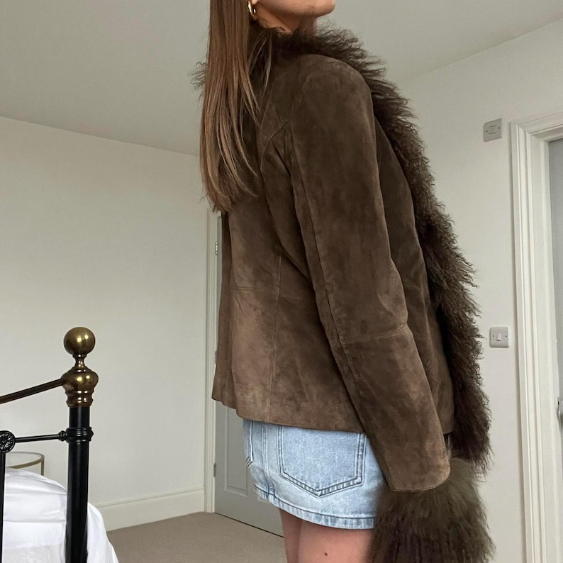 Puede incluir: Chaqueta de ante marr&oacute;n con ribete de piel sint&eacute;tica alrededor del cuello y los pu&ntilde;os. La chaqueta tiene un dise&ntilde;o cl&aacute;sico y una textura suave. La modelo lleva una falda vaquera. Una opci&oacute;n de abrigo elegante.