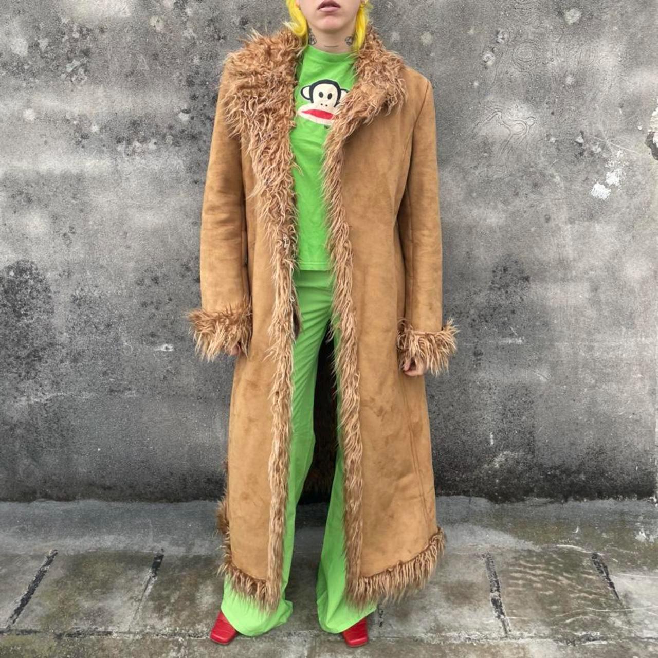 May include: A long, brown faux suede coat with faux fur trim. The coat is worn over a green t-shirt with a monkey graphic and green pants. Red shoes complete the outfit. The coat has a long, flowing design.