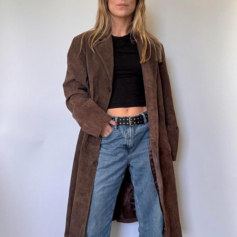 May include: A long, brown suede coat with a button fastening. The coat is worn over a black top and blue jeans. The coat has a classic, tailored design.