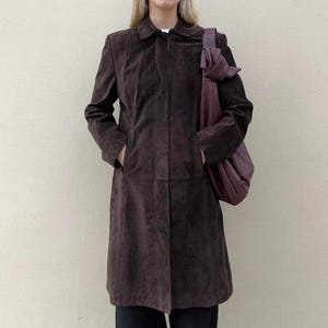 May include: A dark brown suede coat with a collared neckline and pockets. A matching dark purple shoulder bag is worn over the shoulder. The coat is knee-length and has a classic, tailored design.