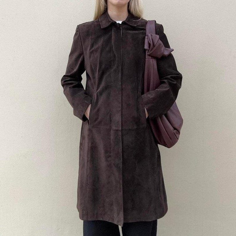 May include: A dark brown suede coat with a collared neckline and pockets. A matching dark purple shoulder bag is worn over the shoulder. The coat is knee-length and has a classic, tailored design.