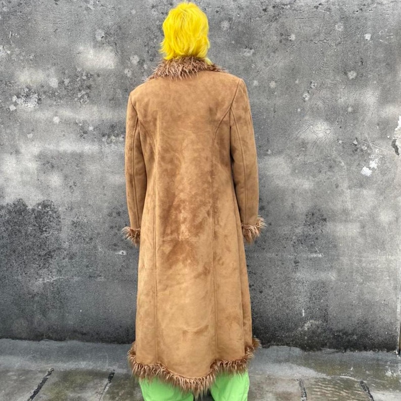 May include: A long, brown suede coat with faux fur trim around the collar, cuffs, and hem. The coat is knee-length and has a classic, tailored design. The person is wearing green pants and has yellow hair.