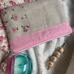 Peut inclure: Une pochette beige et rose à imprimé floral avec un panneau inférieur rose et une fermeture éclair argentée. Un flacon de parfum transparent et rose est dans le coin inférieur gauche. Un charm en perles de bois avec du texte français est suspendu à la fermeture éclair.