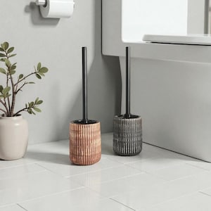 May include: Two toilet brushes with black handles and textured bases. One base is copper-coloured, the other is grey. The brushes are on a white tiled floor near a toilet and a roll of toilet paper.