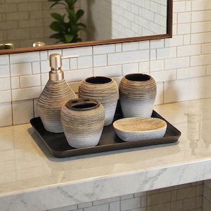 Puede incluir: Un juego de accesorios de baño que incluye un dispensador de jabón, un porta cepillos de dientes, dos vasos y una jabonera. El juego tiene un diseño texturizado a rayas en tonos marrones y blancos, y está sobre una bandeja negra. El dispensador tiene una bomba dorada.
