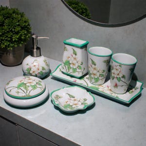 K&ouml;nnte beinhalten: Ein wei&szlig;es Badezimmer-Set aus Keramik mit floralem Design. Das Set umfasst einen Seifenspender, zwei Becher, einen Zahnb&uuml;rstenhalter, eine Seifenschale, ein abgedecktes Glas und zwei Tabletts. Jedes St&uuml;ck hat einen gr&uuml;nen Rand und ein Blumenmuster.