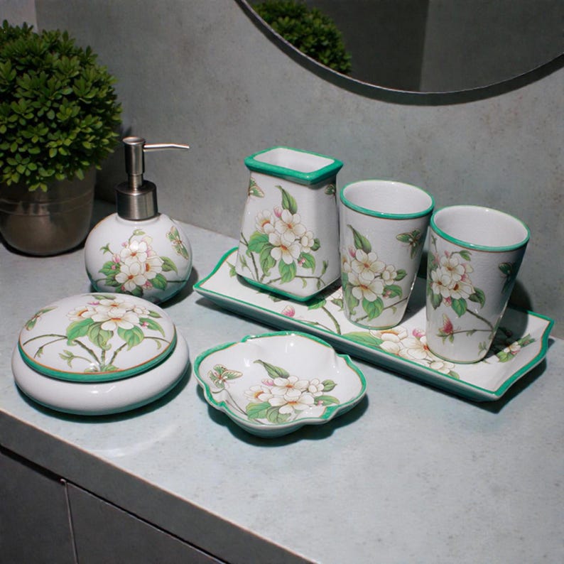 K&ouml;nnte beinhalten: Ein wei&szlig;es Badezimmer-Set aus Keramik mit floralem Design. Das Set umfasst einen Seifenspender, zwei Becher, einen Zahnb&uuml;rstenhalter, eine Seifenschale, ein abgedecktes Glas und zwei Tabletts. Jedes St&uuml;ck hat einen gr&uuml;nen Rand und ein Blumenmuster.
