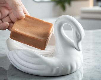 Puede incluir: Un jabonera de cerámica blanca con forma de cisne que sostiene una pastilla de jabón marrón. El cisne tiene un cuello curvo. La jabonera está sobre una superficie de mármol.