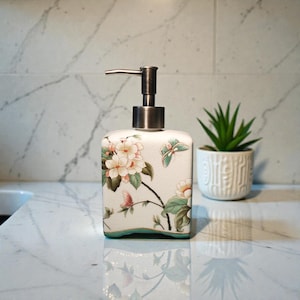 K&ouml;nnte beinhalten: Ein quadratischer Seifenspender aus Keramik mit Blumen- und Schmetterlingsmuster. Er hat eine Pumpe aus geb&uuml;rstetem Metall und einen cremefarbenen Sockel mit t&uuml;rkisfarbenem Rand. Das Blumendesign zeigt rosa und wei&szlig;e Bl&uuml;ten.