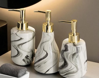 May include: Three white and grey marble-effect soap dispensers with gold-coloured pump tops. A small, folded grey towel sits in a white rectangular tray in front of the dispensers. The dispensers have different shapes.