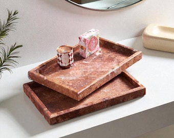 May include: Two rectangular, reddish-brown marble trays stacked on a white surface. A small candle in a patterned holder and a floral-patterned box sit on the top tray. A mirror is in the background.