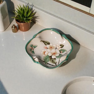 K&ouml;nnte beinhalten: Eine dekorative, wei&szlig;e Keramikschale mit gewelltem Rand und t&uuml;rkisfarbenem Rand. Die Schale zeigt ein Blumendesign mit wei&szlig;en Bl&uuml;ten, gr&uuml;nen Bl&auml;ttern und einem Schmetterling. Die Schale dient wahrscheinlich dekorativen Zwecken.