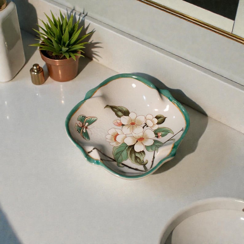 K&ouml;nnte beinhalten: Eine dekorative, wei&szlig;e Keramikschale mit gewelltem Rand und t&uuml;rkisfarbenem Rand. Die Schale zeigt ein Blumendesign mit wei&szlig;en Bl&uuml;ten, gr&uuml;nen Bl&auml;ttern und einem Schmetterling. Die Schale dient wahrscheinlich dekorativen Zwecken.