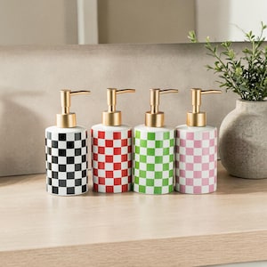 May include: Four ceramic soap dispensers with gold-coloured pumps. Each dispenser has a white base and a chequered pattern in black, red, green, and pink. The dispensers are arranged on a wooden surface.
