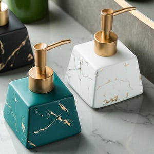 Pode incluir: Dispensadores de sabonete quadrados em cerâmica em tons de verde-azulado, branco e preto, com bombas douradas. Os dispensadores têm um padrão semelhante ao mármore com veios dourados. O design é moderno e adequado para uma casa de banho ou cozinha.