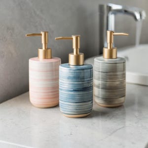 Könnte beinhalten: Drei Keramik-Seifenspender mit goldfarbenen Pumpen. Die Spender sind zylindrisch mit einem gestreiften Muster in Rosa, Blau und Grau. Die Spender stehen auf einer weißen Arbeitsplatte.
