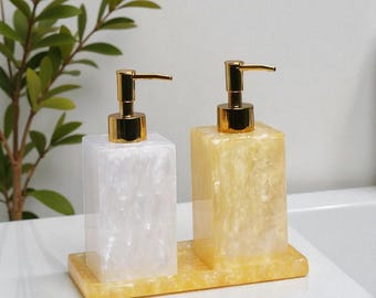 May include: Two decorative soap dispensers with gold-colored pumps. One dispenser is white with a pearlescent effect, and the other is yellow with a similar effect. Both sit on a matching yellow tray, ideal for a bathroom or kitchen.