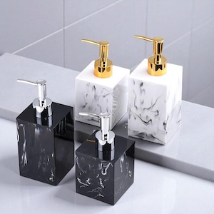 May include: Four square soap dispensers with marble-like patterns. Two dispensers have gold-colored pumps, and two have silver-colored pumps. The dispensers are black and white, and are arranged on a light gray surface.