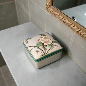 K&ouml;nnte beinhalten: Quadratische, cremefarbene Keramik-Schmuckschatulle mit floralem Design auf dem Deckel. Der Deckel zeigt wei&szlig;e Bl&uuml;ten, gr&uuml;ne Bl&auml;tter und einen Schmetterling. Die Box hat einen gr&uuml;nen Rand.