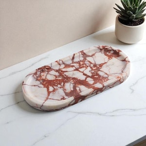 Könnte beinhalten: Ovale Marmorschale mit weißem und rot geädertem Muster. Die Schale hat eine hellrosa Farbe und abgerundete Kanten. Eine kleine Sukkulentenpflanze in einem weißen Topf befindet sich im Hintergrund.