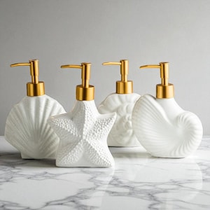 May include: Four white ceramic soap dispensers with gold-coloured pump tops. The dispensers are shaped like seashells and a starfish. The dispensers are arranged on a marble surface.