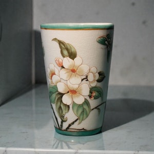K&ouml;nnte beinhalten: Ein wei&szlig;er Keramikbecher mit gr&uuml;nem Rand und Boden, verziert mit einem Blumendesign. Die Bl&uuml;ten sind wei&szlig; mit orangefarbenen Zentren und die Bl&auml;tter sind gr&uuml;n. Der Becher ist etwa 13 cm hoch.