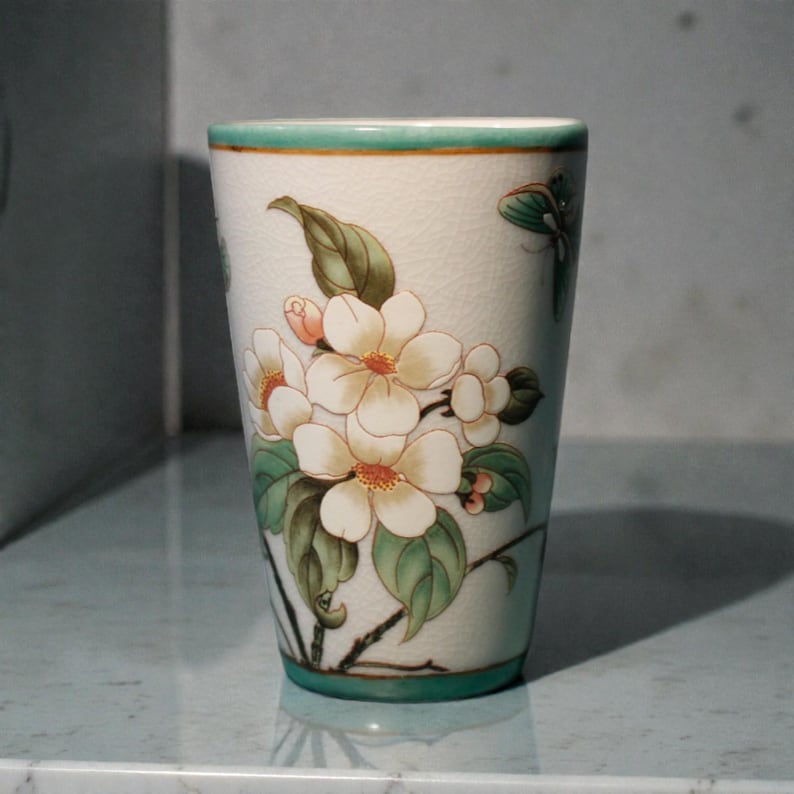 K&ouml;nnte beinhalten: Ein wei&szlig;er Keramikbecher mit gr&uuml;nem Rand und Boden, verziert mit einem Blumendesign. Die Bl&uuml;ten sind wei&szlig; mit orangefarbenen Zentren und die Bl&auml;tter sind gr&uuml;n. Der Becher ist etwa 13 cm hoch.