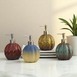 May include: Four ceramic soap dispensers with silver pumps. The dispensers are round with a ribbed design and come in shades of red, blue, yellow, and green. A houseplant in a white pot is in the background.