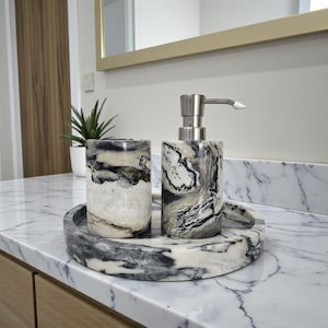 May include: A marble bathroom set featuring a soap dispenser, tumbler, and tray. The set has a swirling pattern of black, white, and gray. The dispenser has a silver pump. The set is on a marble countertop.