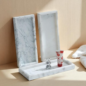 Könnte beinhalten: Drei rechteckige Marmortabletts mit weißem und grau geädertem Muster. Eine kleine silberne Hasenfigur und eine rot-weiße Tube mit Ahornblattmuster befinden sich auf einem Tablett. Die Tabletts stehen auf einer hellen Holzoberfläche.