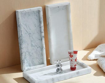May include: Three rectangular marble trays with a white and gray veined pattern. A small silver bunny figurine and a red and white tube with a maple leaf design are on one tray. The trays are on a light wood surface.