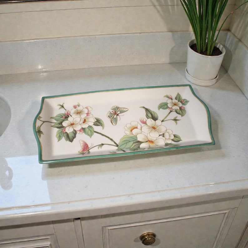 K&ouml;nnte beinhalten: Ein rechteckiges wei&szlig;es Keramiktablett mit Blumen- und Schmetterlingsdesign. Das Tablett hat einen gr&uuml;nen Rand und gebogene Kanten. Die Blumen sind wei&szlig; und rosa, mit gr&uuml;nen Bl&auml;ttern. Die Schmetterlinge sind rosa und gr&uuml;n.