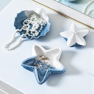 May include: A collection of ceramic trinket dishes in the shape of a seashell and starfish. The seashell dish is blue and white and holds a pearl necklace. The starfish dishes are white and blue and hold jewelry.