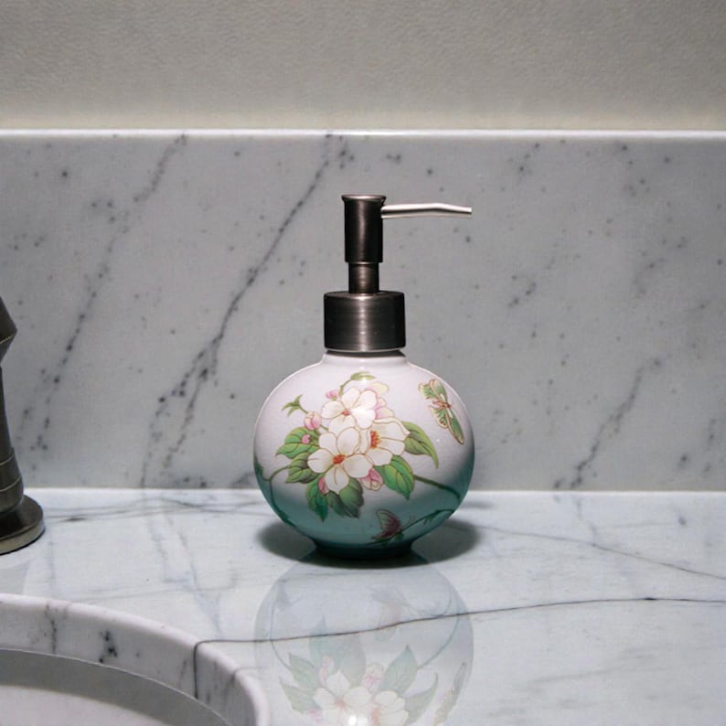 K&ouml;nnte beinhalten: Ein wei&szlig;er Keramik-Seifenspender mit einem Blumendesign aus wei&szlig;en und rosa Bl&uuml;ten und gr&uuml;nen Bl&auml;ttern. Er hat eine Pumpe aus geb&uuml;rstetem Metall und steht auf einer Marmor-Arbeitsplatte. Das Design ist elegant und dekorativ.