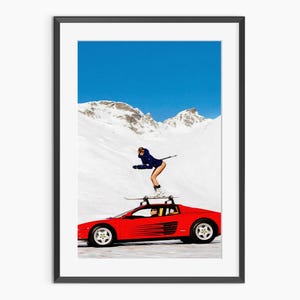 May include: Framed art print showing a red sports car with skis on its roof, set against a snowy mountain and a blue sky. A person in a blue ski suit is skiing on top of the car.